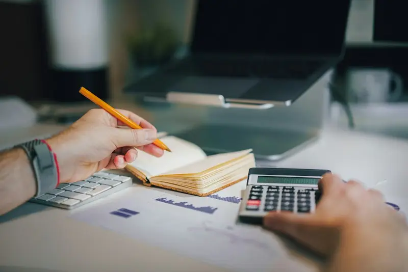 Mãos fazendo contas na calculadora ao lado de relatórios e um caderno